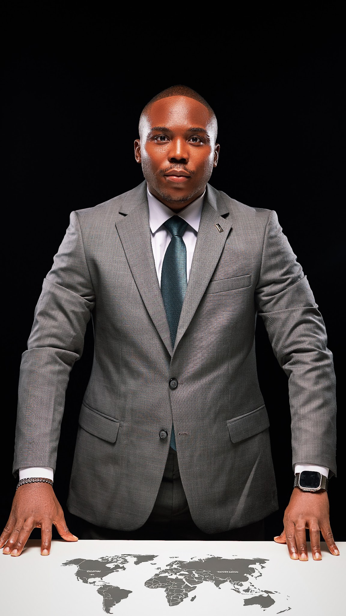 Ekulide Miracle in a grey suit, standing at a table displaying a world map against a dark studio background