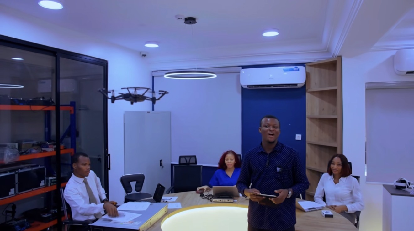 A speaker addresses colleagues seated around the lit table in the Miva Open University Robotics & Innovation Laboratory, with a drone and equipment shelves behind